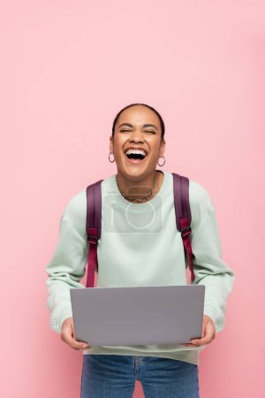 heureux étudiant afro-américain avec sac à dos tenant ordinateur portable et riant isolé sur rose 
