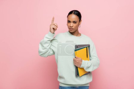 smart african american student holding laptop and notebooks while having idea isolated on pink 