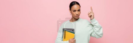 étudiant afro-américain intelligent tenant ordinateur portable et fournitures d'étude tout en ayant idée isolé sur rose, bannière 