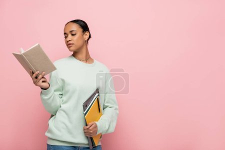 smart african american student in sweatshirt reading book while holding laptop and notebooks isolated on pink 