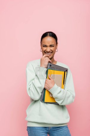 cheerful african american student in sweatshirt holding laptop and study supplies isolated on pink 