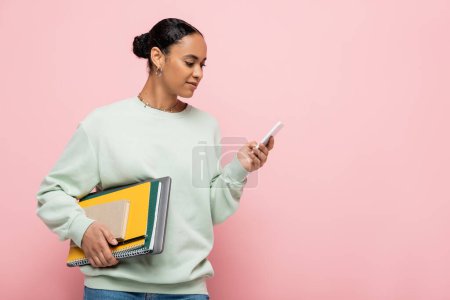 pretty african american student in sweatshirt holding study supplies and using smartphone isolated on pink 
