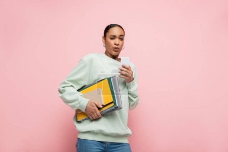 Angespannte afrikanisch-amerikanische Studentin in Sweatshirt mit Studienmaterial und Smartphone isoliert auf rosa 