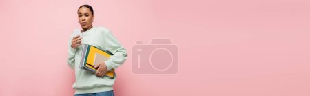 tensed african american student in sweatshirt holding study supplies and using smartphone isolated on pink, banner 