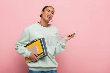 displeased african american student in sweatshirt holding gadgets and study supplies while whining isolated on pink 