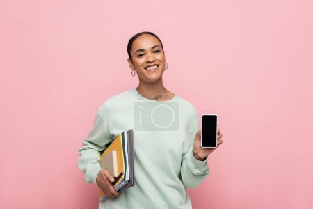 lächelnde afrikanisch-amerikanische Studentin im Sweatshirt mit Lernmaterial und Smartphone mit leerem Bildschirm, isoliert auf rosa 