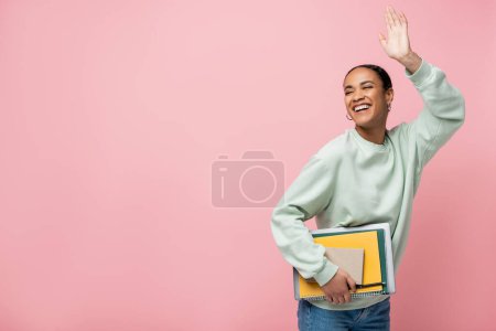 fröhliche afrikanisch-amerikanische Studentin in Sweatshirt mit Studienmaterial und winkender Hand isoliert auf rosa 