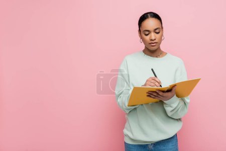 pretty african american student in sweatshirt taking notes while studying isolated on pink 