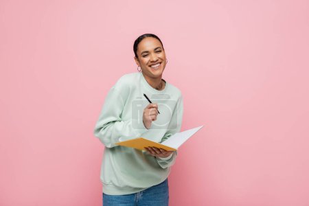 cheerful african american student in sweatshirt holding pen and notebook isolated on pink 
