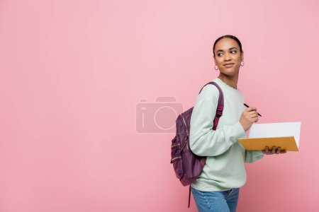 lächelnder afrikanisch-amerikanischer Student mit Rucksack, Stift und Notizbuch isoliert auf rosa 