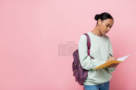 brunette african american student taking notes while holding notebook and pen isolated on pink 