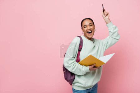 estudiante afroamericano emocionado sosteniendo portátil y pluma mientras está de pie con mochila aislada en rosa 