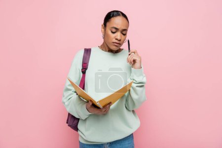 frustrated african american student holding notebook and pen while standing with backpack isolated on pink 