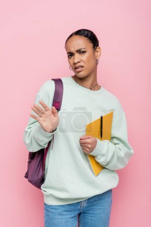 displeased african american student holding notebook and pen while showing refuse gesture isolated on pink 