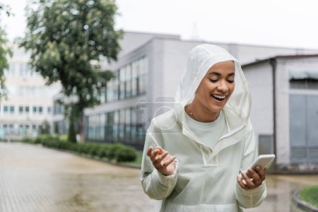 excited african american woman in waterproof raincoat with hood looking at smartphone while standing under rain 