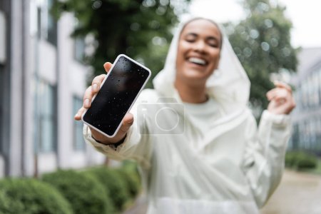 alegre mujer afroamericana en impermeable con capucha sosteniendo teléfono inteligente húmedo con pantalla en blanco 