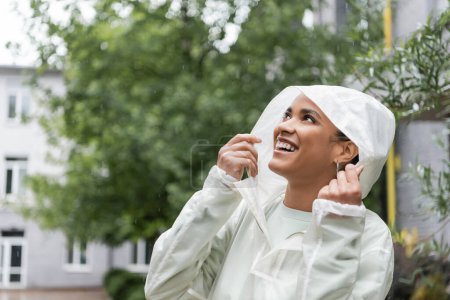 overjoyed african american woman in waterproof raincoat with hood looking up during rain 