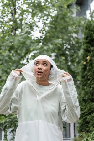 surprised african american woman in waterproof raincoat and wireless headphones standing under rain 