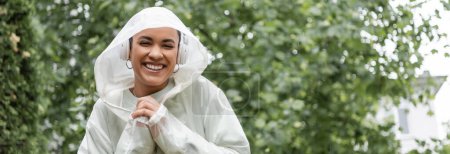 sonriente mujer afroamericana en impermeable y auriculares inalámbricos de pie bajo la lluvia, bandera 
