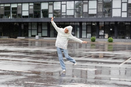 volle Länge der glücklichen afrikanisch-amerikanischen Frau in wasserdichtem Regenmantel und Jeans, die Spaß bei Regen hat 