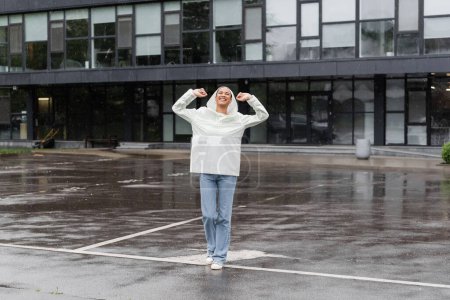 Téléchargez les photos : Pleine longueur de femme afro-américaine ravie en imperméable imperméable et jeans s'amuser pendant la pluie - en image libre de droit