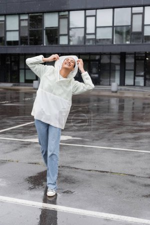 Foto de Longitud completa de la mujer afroamericana complacida en impermeable y jeans que se divierten durante la lluvia - Imagen libre de derechos