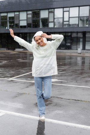 Foto de Longitud completa de la mujer afroamericana positiva en impermeable y pantalones vaqueros divertirse durante la lluvia - Imagen libre de derechos