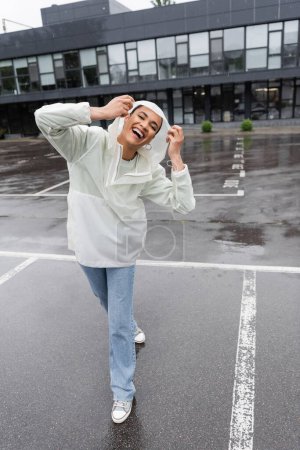 volle Länge der aufgeregten afrikanisch-amerikanischen Frau in wasserdichtem Regenmantel und Jeans, die Spaß bei Regen hat 