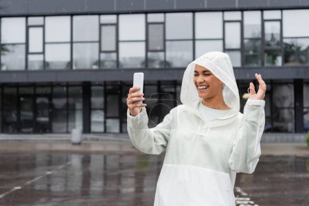 Téléchargez les photos : Heureuse femme afro-américaine en imperméable à l'eau prenant selfie sur smartphone pendant la pluie - en image libre de droit