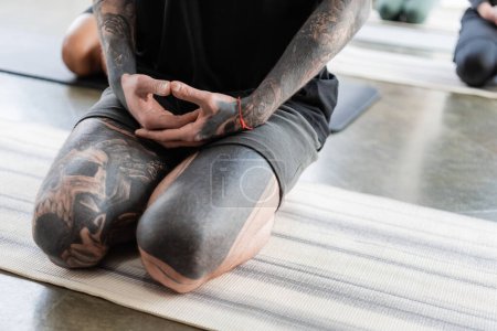 Cropped view of tattooed man sitting in Thunderbolt asana on yoga mat in studio 