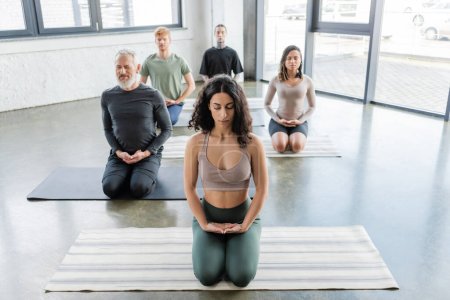 Personas multiétnicas meditando con los ojos cerrados en Thunderbolt asana en estudio de yoga 