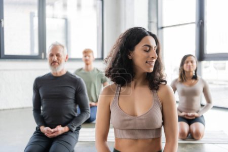 Sonriente mujer de Oriente Medio meditando cerca de personas borrosas en clase de yoga 