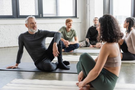 Hombre maduro positivo hablando con la mujer de Oriente Medio en clase de yoga 