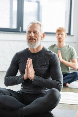 Homme mûr méditant avec les yeux fermés et anjali mudra sur tapis en cours de yoga 