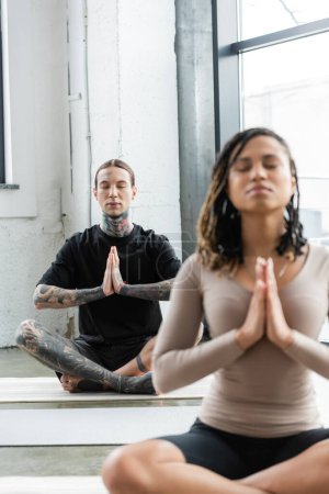 Joven hombre tatuado meditando y haciendo anjali mudra en clase de yoga 