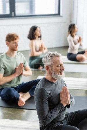 Mature man sitting in anjali mudra near blurred multiethnic group in yoga class 