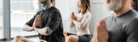 Homme tatoué faisant anjali mudra près de groupe interracial en cours de yoga, bannière 