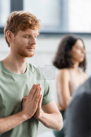 Redhead man with closed eyes doing anjali mudra in yoga class 