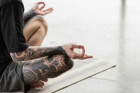 Téléchargez les photos : Vue recadrée d'un jeune homme tatoué pratiquant le gyan mudra sur tapis dans un studio de yoga - en image libre de droit