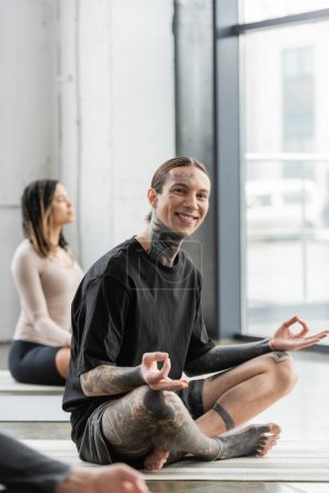 Photo for Positive tattooed man looking at camera while doing gyan mudra in yoga studio - Royalty Free Image