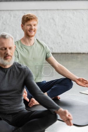 Foto de Alegre pelirroja hombre haciendo gyan mudra y mirando cámara en yoga studio - Imagen libre de derechos