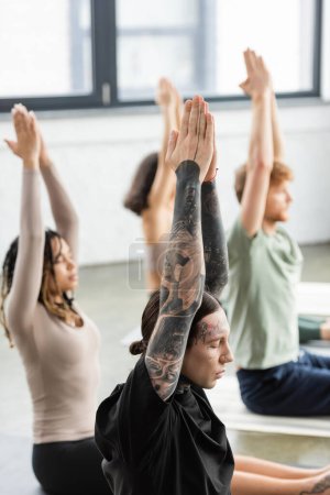 Tattooed man doing anjali mudra while sitting near blurred people in yoga class 