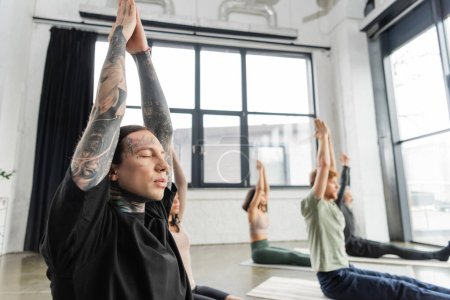Tattooed man doing anjali mudra near blurred group in yoga studio 