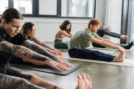 Young african american woman stretching on mat near people in yoga studio 