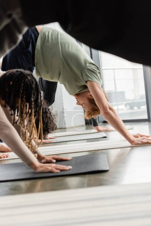 Side view of redhead man standing in Downward Facing Dog asana near interracial group in yoga class