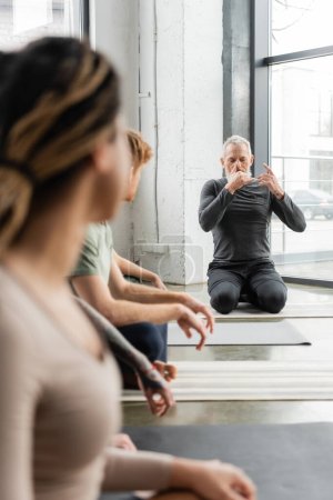 Mature coach practicing nostril breathing near blurred interracial group in yoga class 
