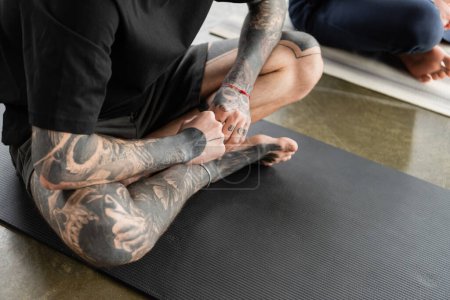 Ausgeschnittene Ansicht eines tätowierten Mannes, der in einem Feuerprotokoll Asana auf einer Matte im Yoga-Kurs sitzt  