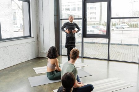 Middle aged coach talking and gesturing near people on mats in yoga class 