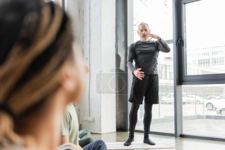 Photo for Mature coach talking and explaining asana to blurred interracial group in yoga class - Royalty Free Image