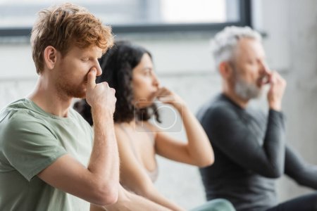 Redhead man practicing nostril breathing near blurred group in yoga studio 